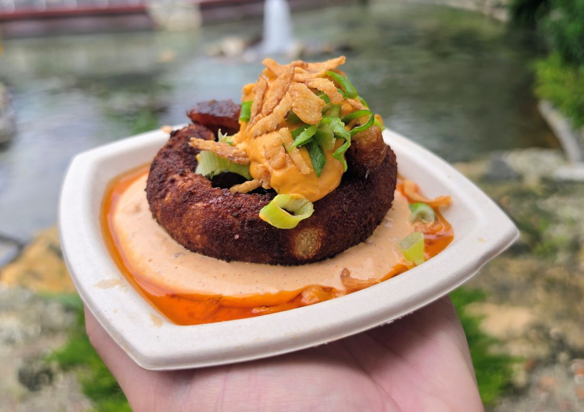 A close-up view of a Loaded Brisket Burnt End Stuffed Potato Ball with chipotle lime crema, beer cheese, crispy onions, and sliced scallions, presented in a square paper dish held over a blurred outdoor setting at Busch Gardens Bier Fest
