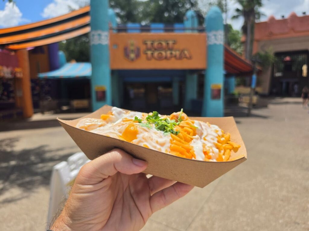 Hand holding a paper food boat of loaded tater tots topped with shredded cheese, white sauce, and cilantro in front of the Tot-Topia dining location.