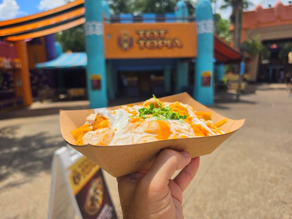 Side angle of loaded tater tots served in a cardboard boat held against a blurred background of the Tot-Topia building.