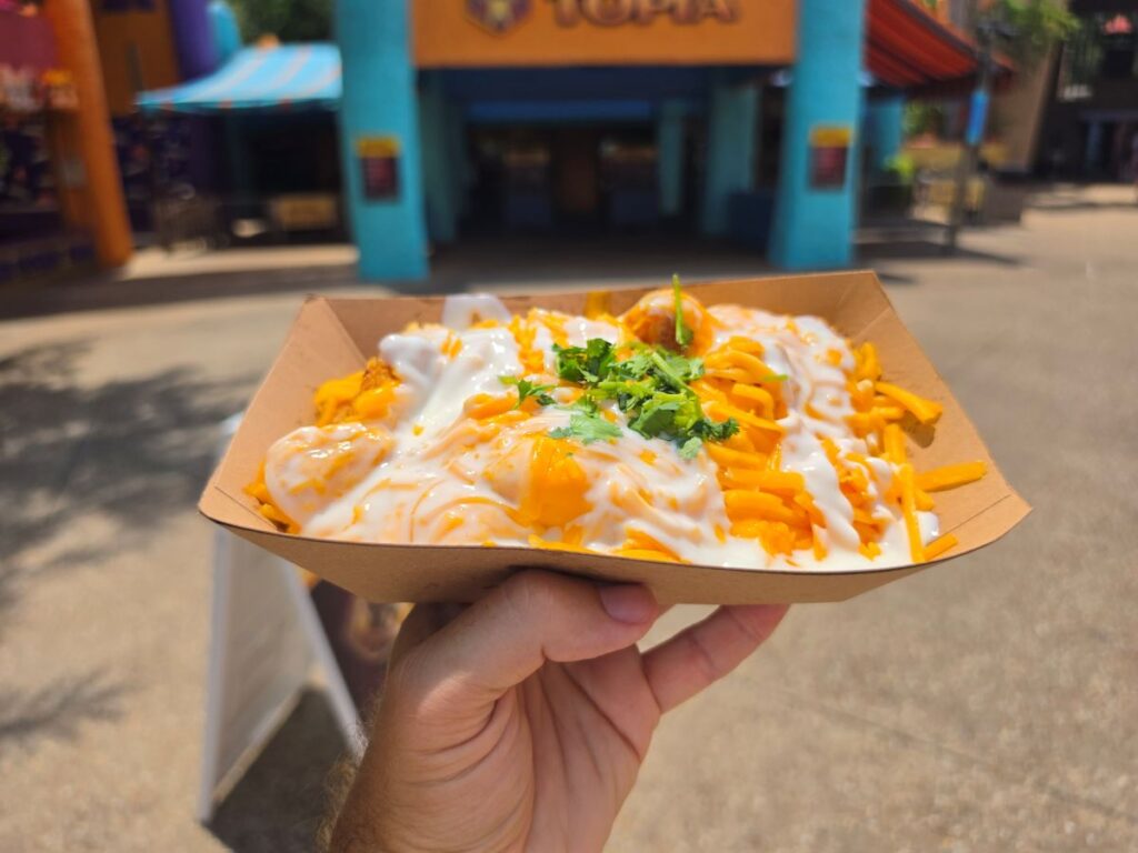Close-up view of a hand holding a paper boat filled with loaded tater tots with melted cheese, drizzle, and herbs .