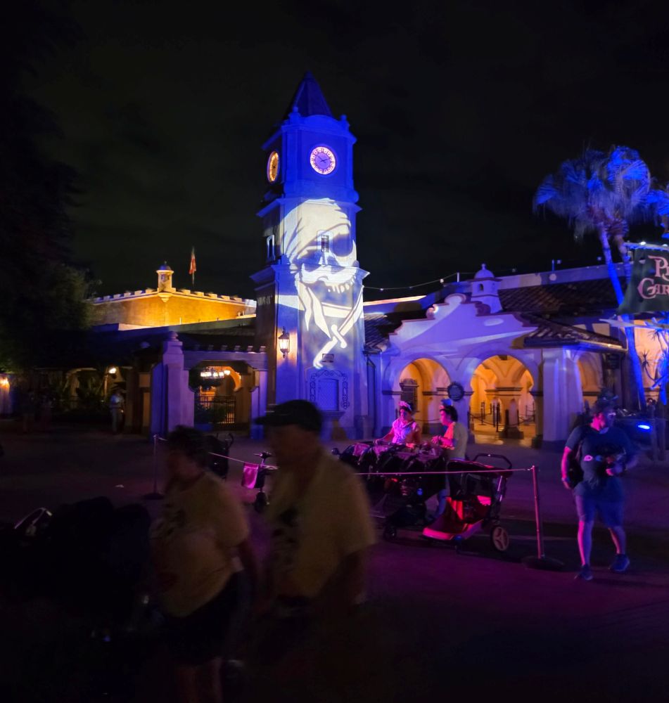 A clock tower at night features a large projected skull and crossbones, with people walking in the foreground of the illuminated theme park area.