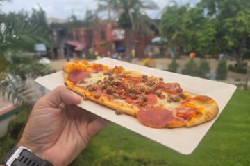 A hand holding an oblong meat lovers flatbread pizza topped with pepperoni, sausage, and bacon on a paper tray, shot from an angle with an outdoor theme park setting in the background.