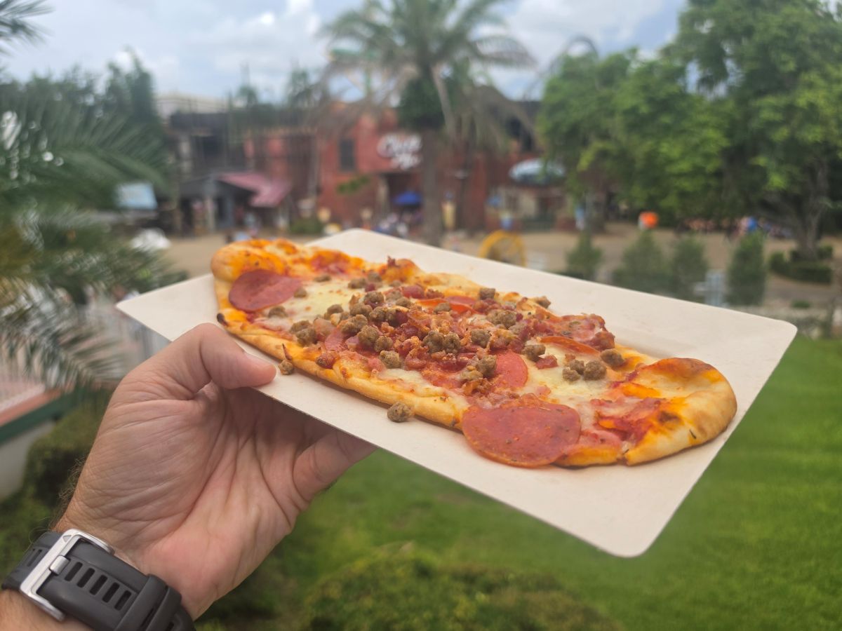 A hand holding an oblong meat lovers flatbread pizza topped with pepperoni, sausage, and bacon on a paper tray, shot from an angle with an outdoor theme park setting in the background.