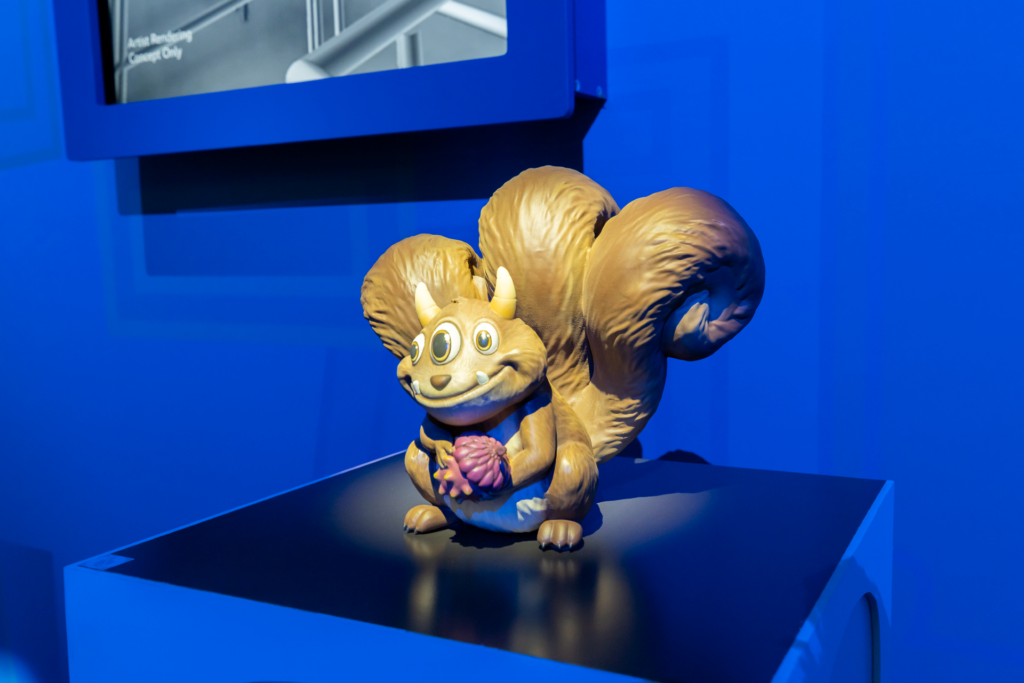 A brown, three-eyed, horned squirrel figurine with a bushy tail holds a small, spiky purple object while sitting on a dark pedestal against blue walls.