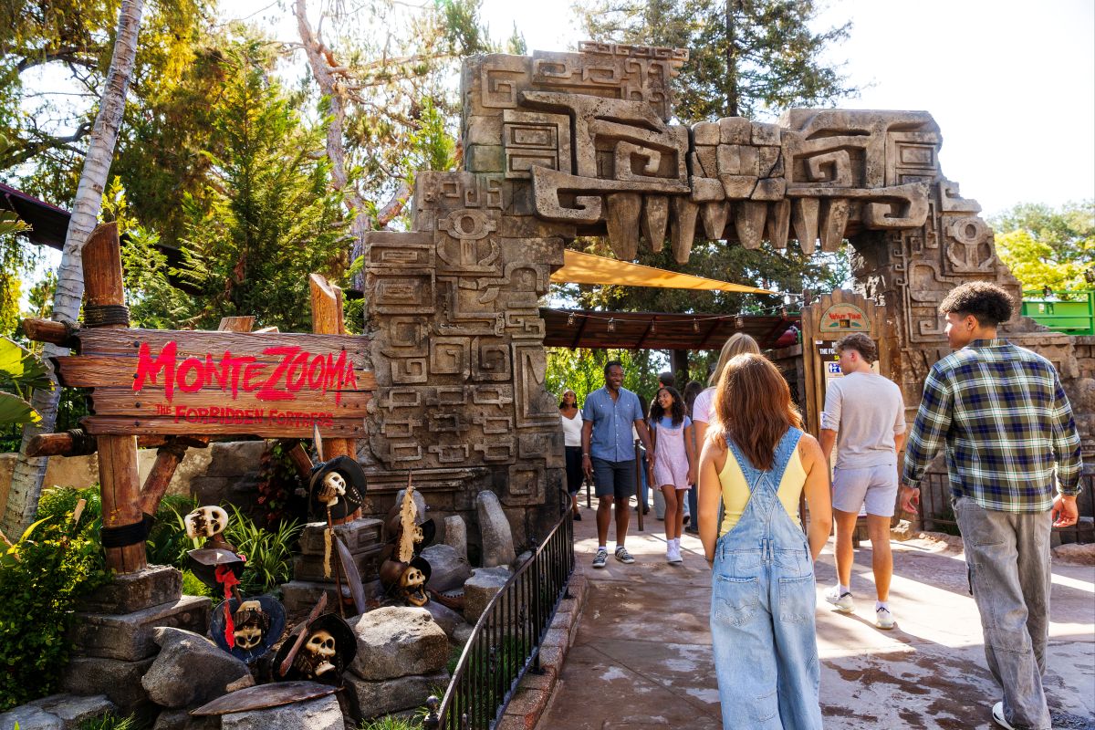 Visitors walk through a stone archway carved with ancient designs at the Montezooma: The Forbidden Fortress attraction entrance at a theme park.