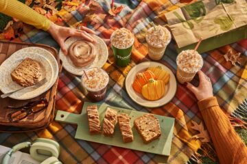 A picnic on a plaid blanket features Panera Fall menu pastries, iced drinks with whipped cream, pumpkin cookies, and sliced loaf cake on a wooden board.