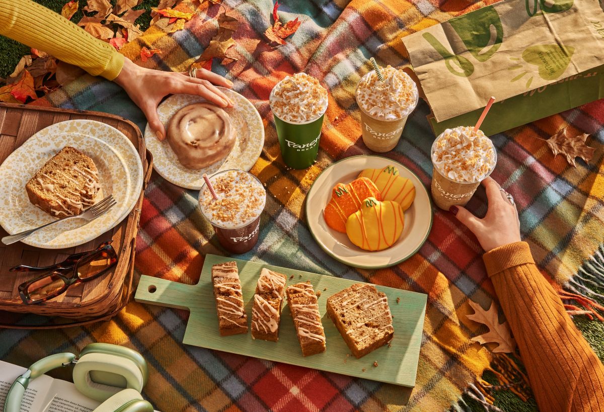 A picnic on a plaid blanket features Panera Fall menu pastries, iced drinks with whipped cream, pumpkin cookies, and sliced loaf cake on a wooden board.