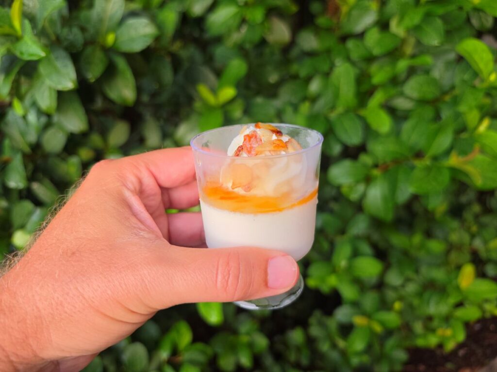 A hand holds a small plastic cup containing a layered dessert with white cream, orange fruit, and sliced almonds, set against green shrubbery.