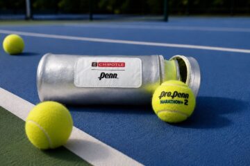 A silver Penn tennis ball canister with a Chipotle logo sits on a blue tennis court next to three yellow tennis balls, one resting near the open lid.