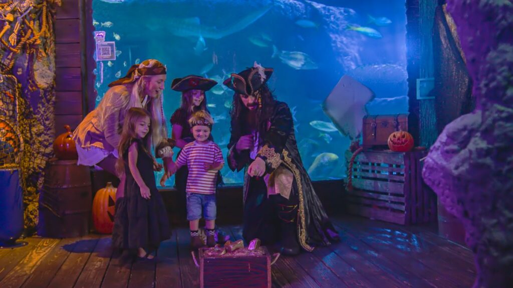 A pirate kneels before children in costumes near a large aquarium, pointing at a small wooden treasure chest filled with gold-like items.