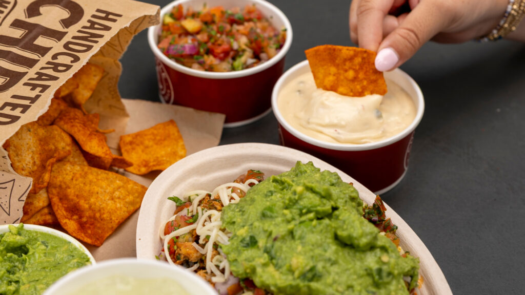 A hand dipping a chili lime tortilla chip into a side of warm queso blanco next to a Chipotle Pollo Asado loaded with guacamole, salsa, and cheese.