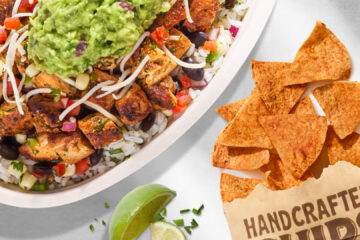 An overhead view of a Chipotle Pollo Asado bowl filled with grilled chicken, rice, black beans, and guacamole alongside seasoned tortilla chips in a brown paper bag and lime wedges.