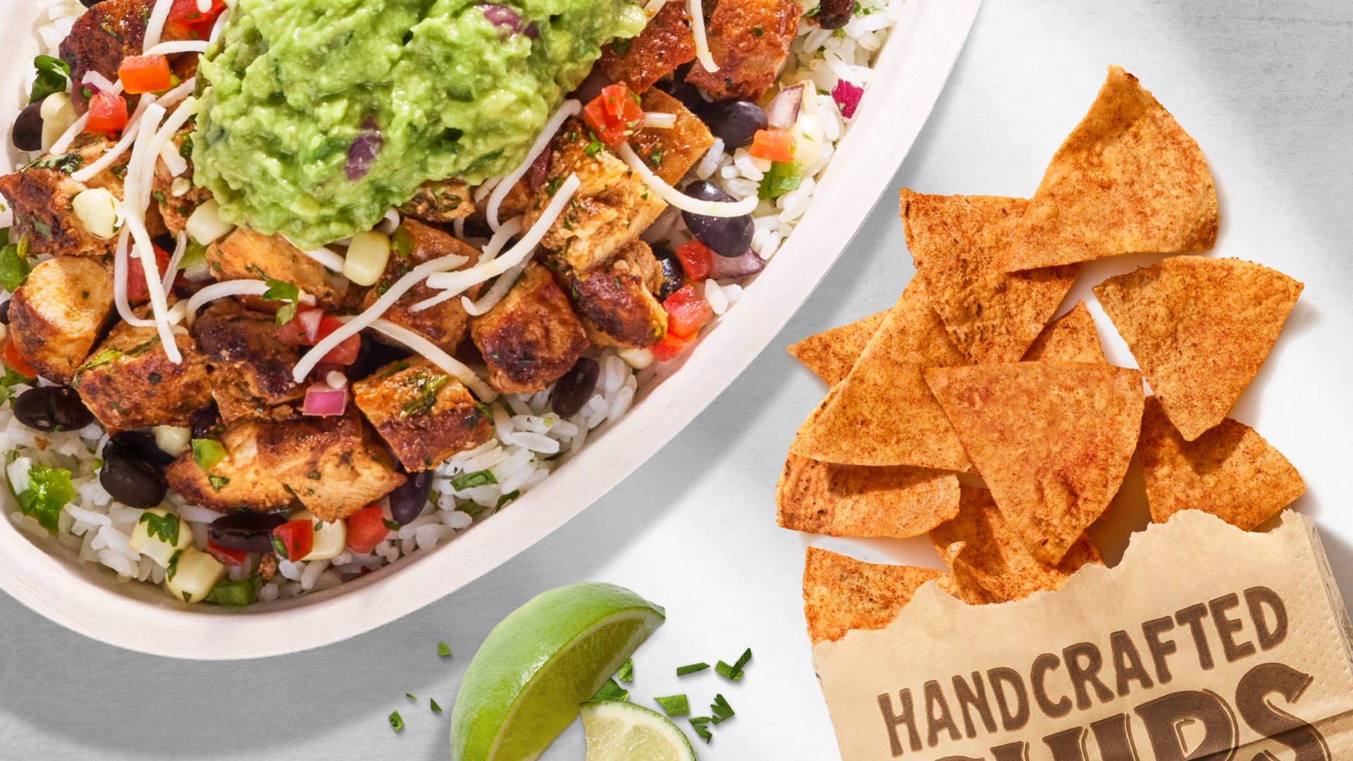 An overhead view of a Chipotle Pollo Asado bowl filled with grilled chicken, rice, black beans, and guacamole alongside seasoned tortilla chips in a brown paper bag and lime wedges.