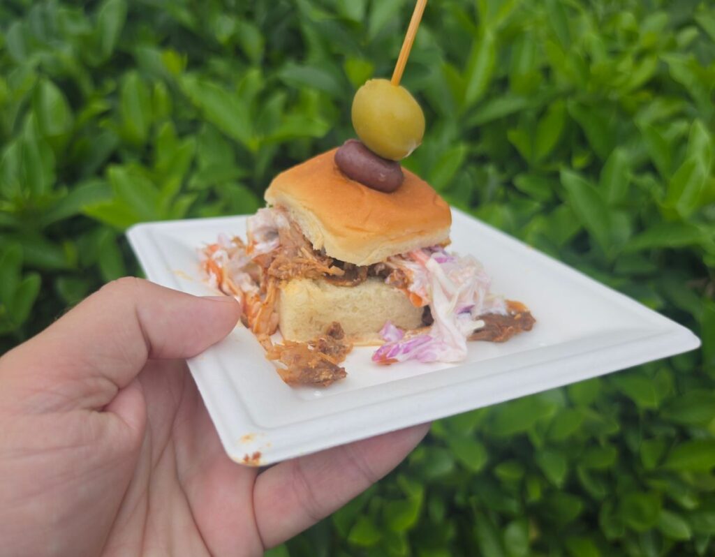 A hand holds a white square plate with a pulled pork slider topped with a green and a black olive on a toothpick, against a blurred green background.