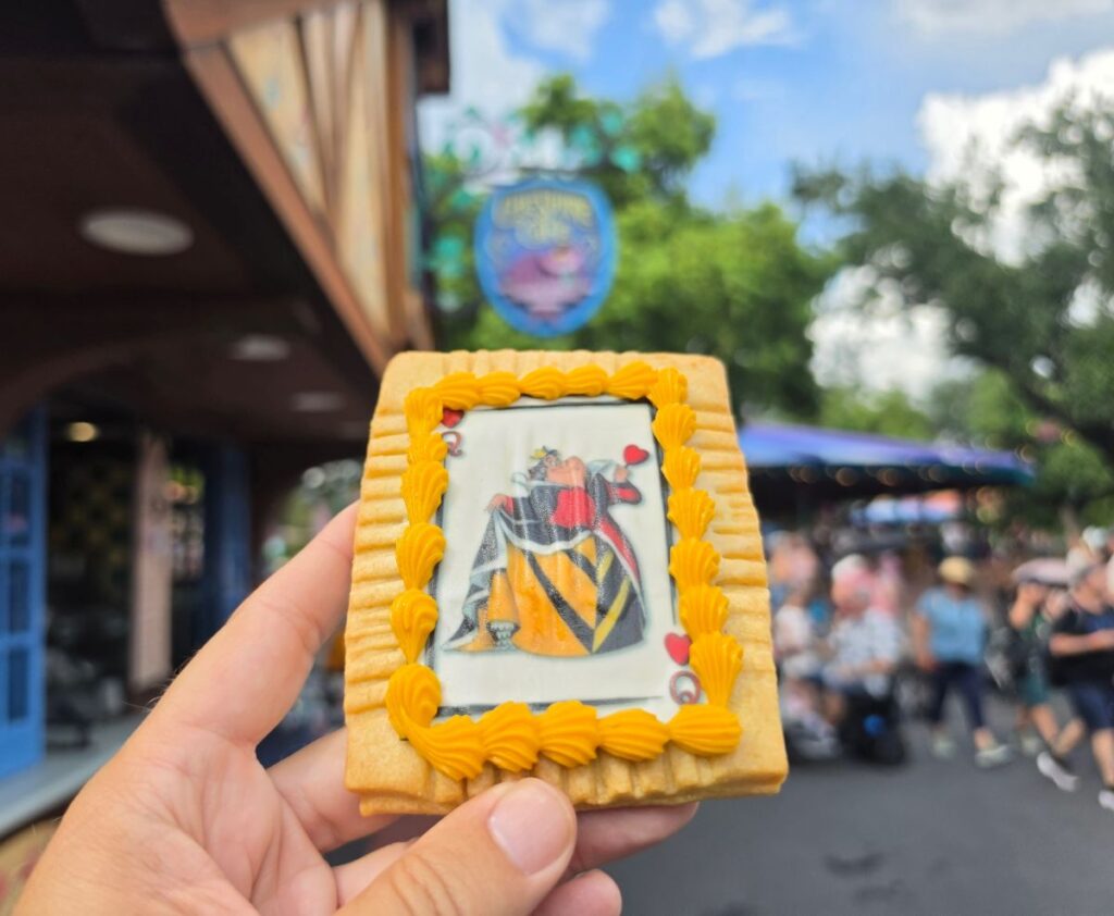 Hand holding a Queen of Hearts Tart with yellow icing trim and a playing card design outside Cheshire Café.