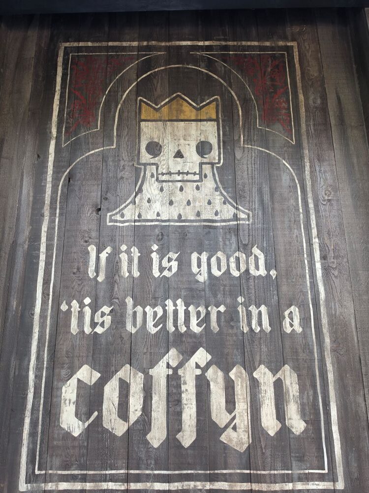 Wooden sign with white Old English text reading "If it is good, 'tis better in a coffyn" below a crowned skull emblem at Six Ravens in Disney Springs.