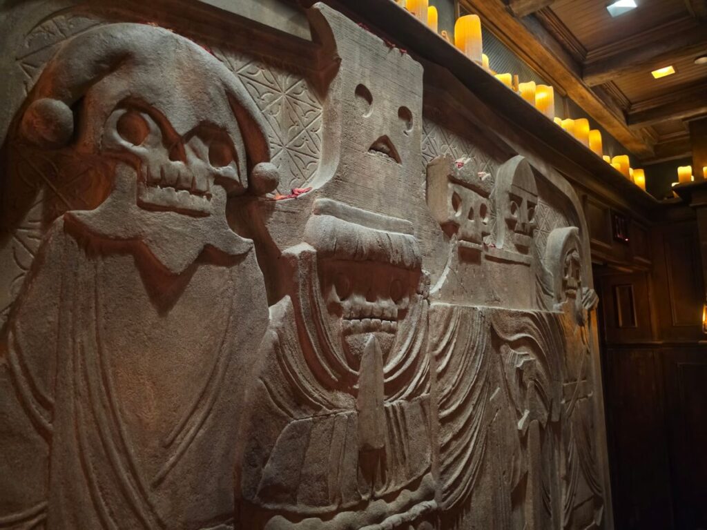 Close-up view of a carved stone wall relief depicting stylized skeleton figures wearing robes and hoods inside Six Ravens at Disney Springs.