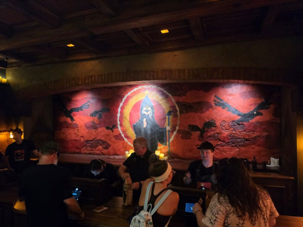 Ordering counter at Six Ravens showing employees serving guests in front of a red medieval wall mural featuring a hooded figure surrounded by ravens.
