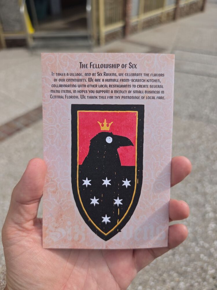 Hand holding the back of the Six Ravens menu, displaying a red and black crest with a crowned raven and six stars alongside "The Fellowship of Six" story text.