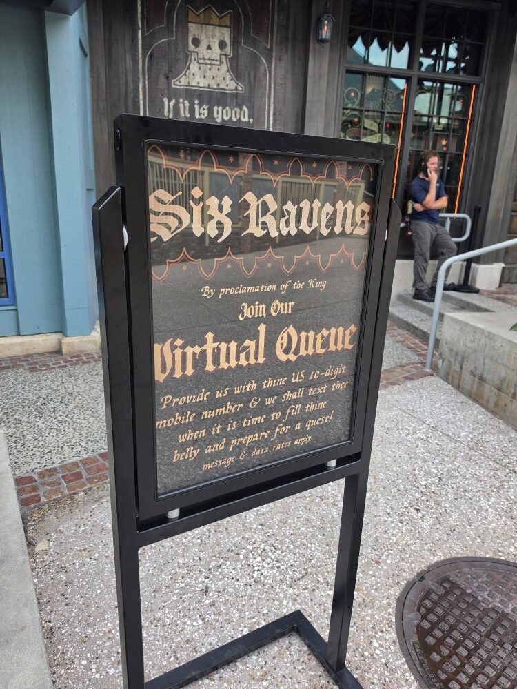 A black framed A-frame sign standing outdoors instructing guests to join the Six Ravens Virtual Queue via mobile phone number.