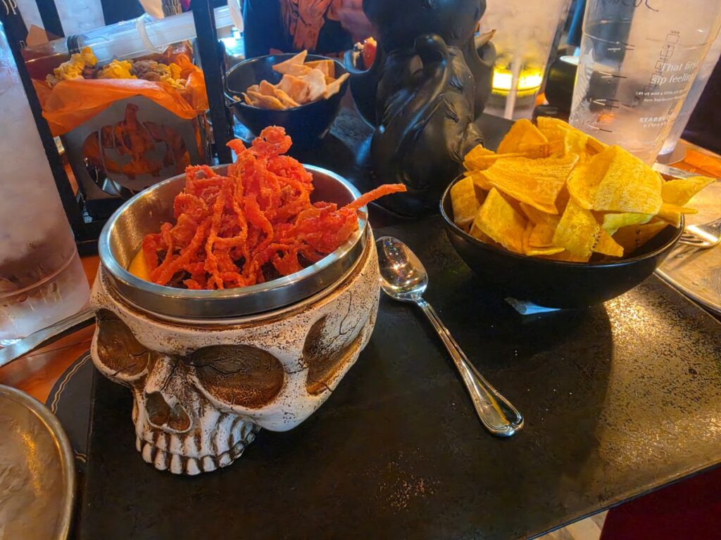 Skull of the Dead Dip topped with crispy red fried onions in a ceramic skull bowl served with a side bowl of plantain chips.