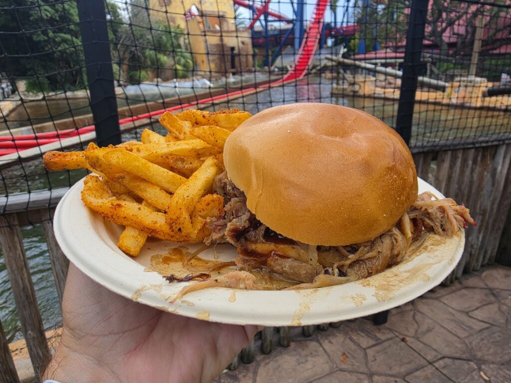 Close-up of a Smoked BBQ Pulled Pork Sandwich dripping with sauce next to seasoned fries outdoors.