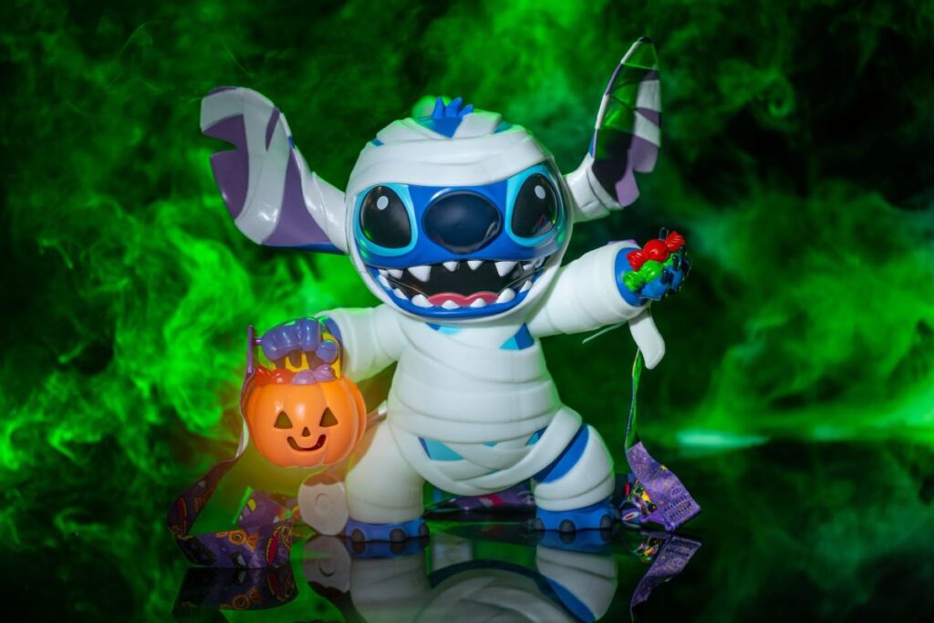 Disney Halloween popcorn bucket featuring Stitch dressed as a mummy holding a glowing jack-o'-lantern trick-or-treat bucket against a green smoky background