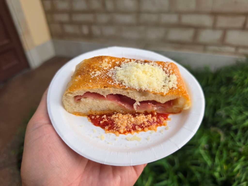 A hand holds a small paper plate with a slice of ham and cheese stuffed bread topped with grated cheese, sitting on a smear of red tomato sauce.
