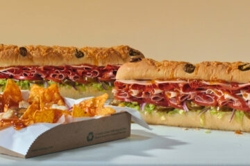 Two long Subway sandwiches with meat, cheese, and vegetables sit next to a cardboard tray filled with orange tortilla chips drizzled with sauce.