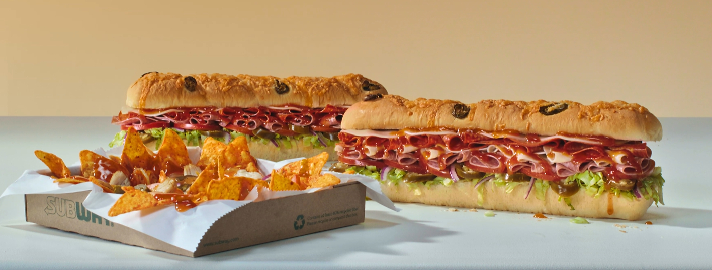 Two long Subway sandwiches with meat, cheese, and vegetables sit next to a cardboard tray filled with orange tortilla chips drizzled with sauce.