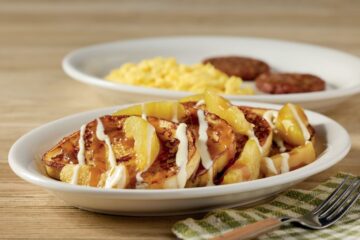 Sweet cream caramel apple French toast topped with sliced apples, drizzled with caramel and sweet cream glaze, served alongside scrambled eggs and sausage patties.