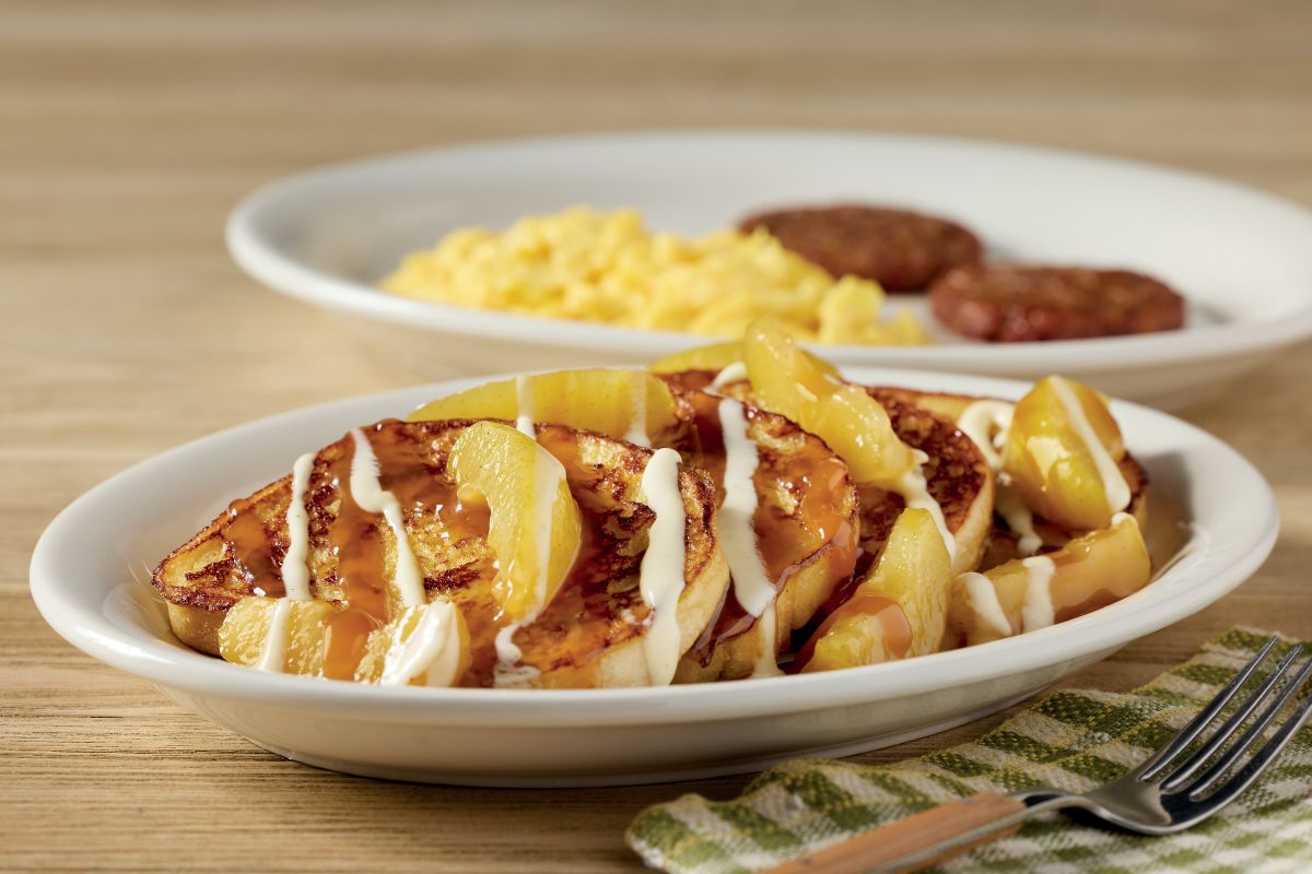 Sweet cream caramel apple French toast topped with sliced apples, drizzled with caramel and sweet cream glaze, served alongside scrambled eggs and sausage patties.