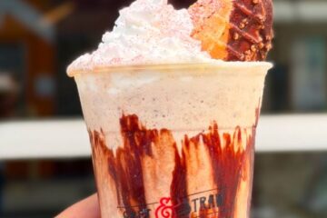 A hand holds a Salt & Straw Tacolate milkshake, drizzled with chocolate syrup and topped with whipped cream and a chocolate-dipped waffle cone piece, in a cup.