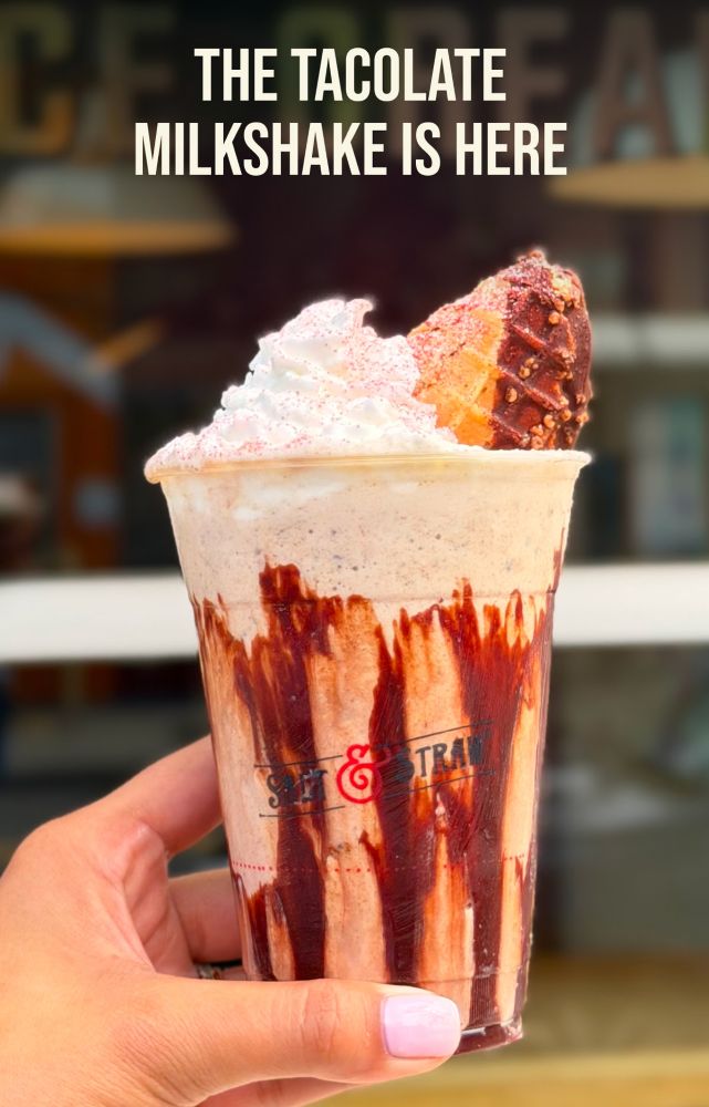 A hand holds a Salt & Straw Tacolate milkshake, drizzled with chocolate syrup and topped with whipped cream and a chocolate-dipped waffle cone piece, in a cup.