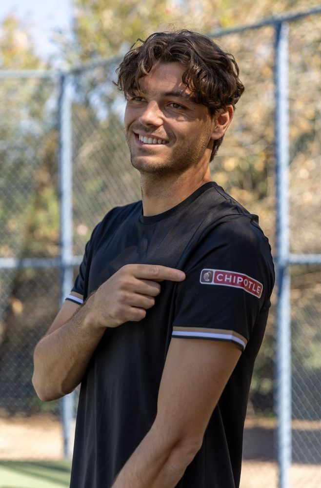 A smiling young man, Taylor Fritz, with wavy brown hair, wears a black T-shirt with a Chipotle logo on the sleeve, pointing toward it while standing near a fence for Chipotle BOGO tennis promotion