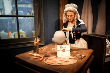 A wax figure of a woman in period clothing works on a blank mannequin head at a wooden table with art supplies, a palette, and a sealed deed for Great Wax Heist
