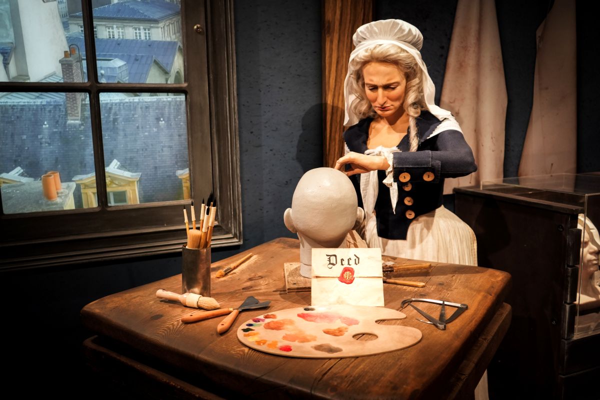 A wax figure of a woman in period clothing works on a blank mannequin head at a wooden table with art supplies, a palette, and a sealed deed for Great Wax Heist