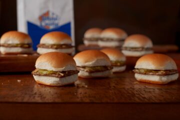 Several White Castle sliders with beef patties, pickles, and onions on soft buns arranged on a wooden surface with a branded bag in the background for White Castle slider deal