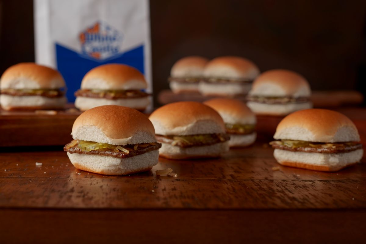 Several White Castle sliders with beef patties, pickles, and onions on soft buns arranged on a wooden surface with a branded bag in the background for White Castle slider deal