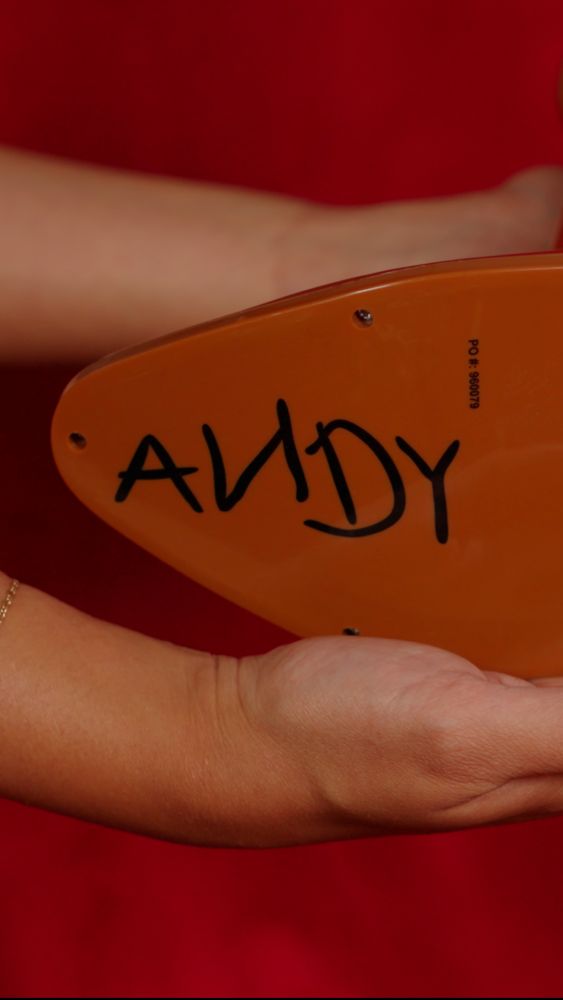A person holds a smooth, orange, pointed object with the name ANDY written in black marker and a small PO number printed on a red background.