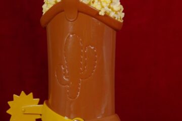 Woody's Boot Popcorn Bucket photo - A hand reaches into a brown plastic cowboy boot container filled with popcorn, featuring a yellow spur, set against a solid dark red background.