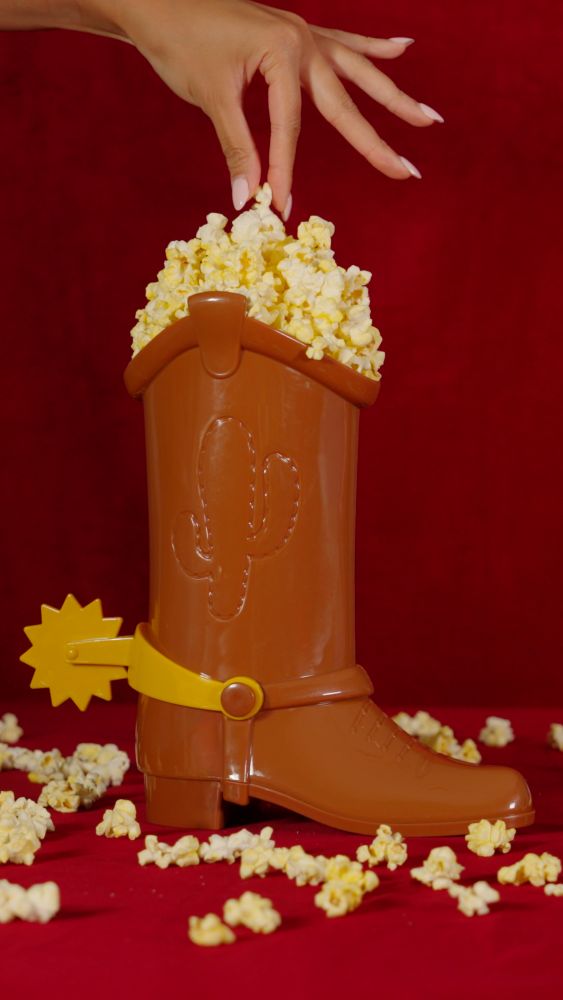 Woody's Boot Popcorn Bucket photo - A hand reaches into a brown plastic cowboy boot container filled with popcorn, featuring a yellow spur, set against a solid dark red background.