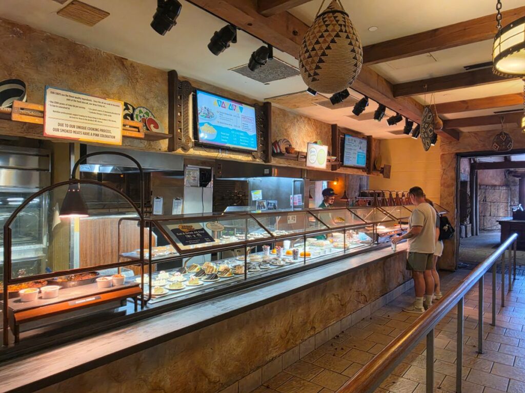 Busch Gardens Tampa Bay quick-service cafeteria counter with display cases featuring desserts and side dishes, plus digital menu screens.