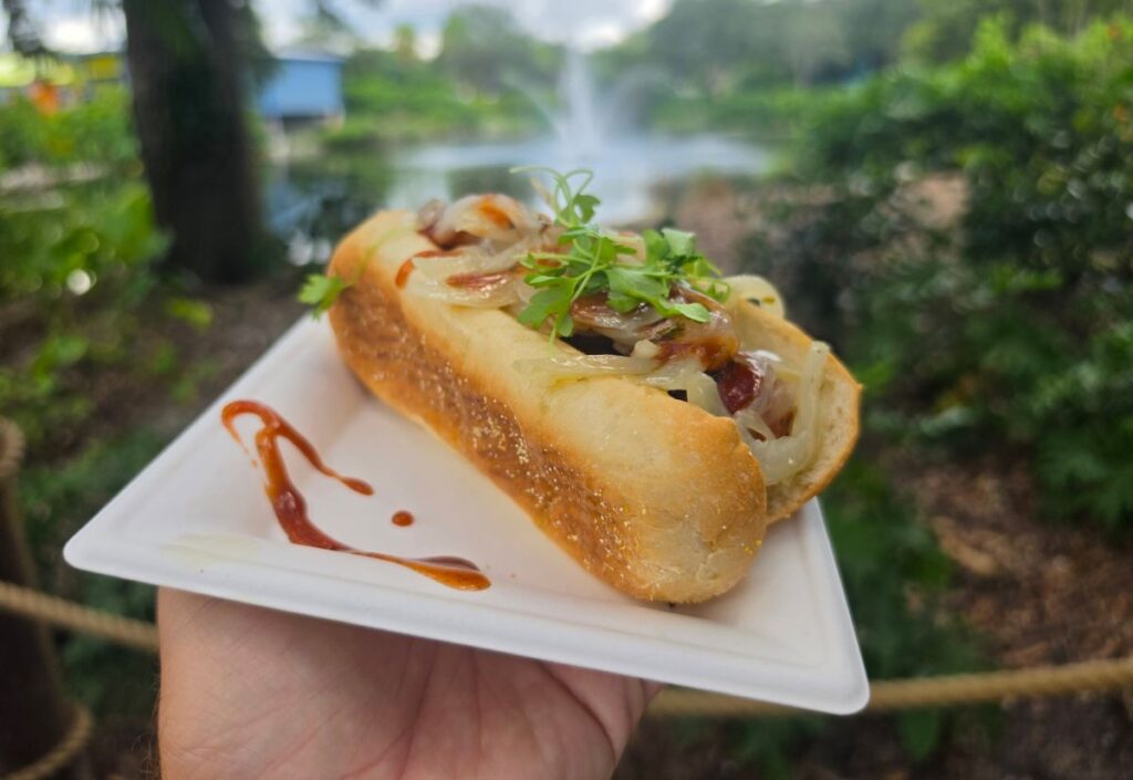 A hand holds a Seaworld Red White BBQ festival white plate with a sausage topped with sautéed onions, sauce, and fresh herbs, set against a blurred park background with a fountain.