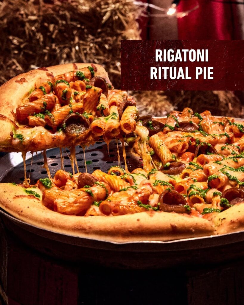 A slice of Rigatoni Ritual Pie lifted from a whole pizza topped with rigatoni pasta, pepperoni, and green pesto drizzle at Halloween Horror Nights 35.