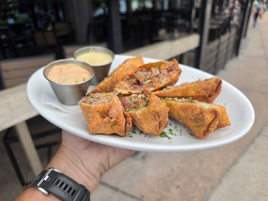 Hand holding a plate of crispy smoked brisket Reuben egg rolls served with dipping sauces outdoors at City Works.