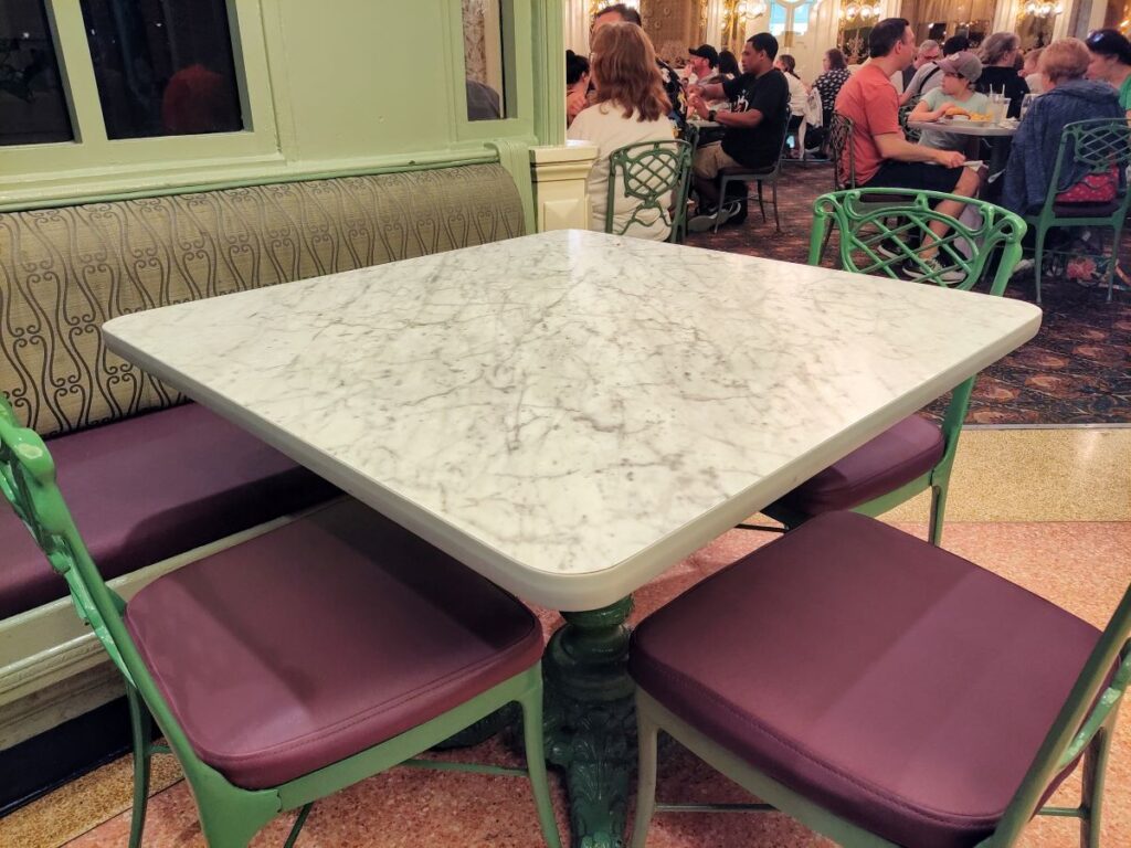 An empty square marble table with green metal chairs and purple cushions inside The Plaza Restaurant at Magic Kingdom.