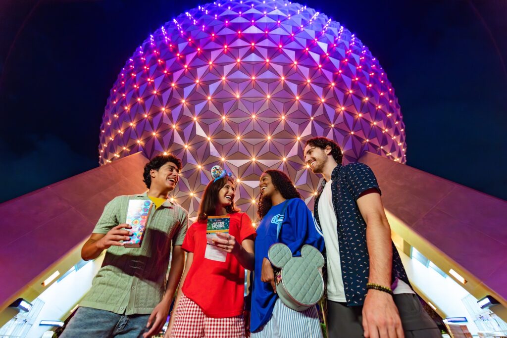 Four friends stand smiling in front of the illuminated Spaceship Earth dome at night, holding park maps and wearing casual clothes,