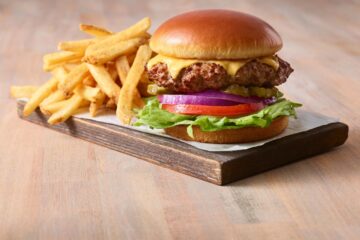 A cheeseburger with lettuce, tomato, onion, and pickles sits on a wooden board next to a pile of golden french fries on a light wood surface.