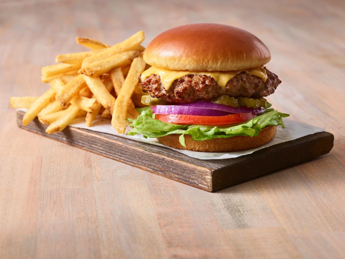 A cheeseburger with lettuce, tomato, onion, and pickles sits on a wooden board next to a pile of golden french fries on a light wood surface.